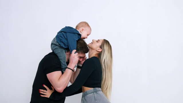 Man holding a little baby on shoulders. Woman tries to kiss her child reaching to him. White backdrop.