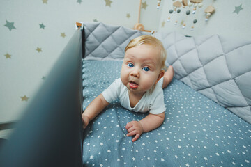 Photo of a 5-month-old baby lying in a crib. A cot with a little boy in a white bodysuit.