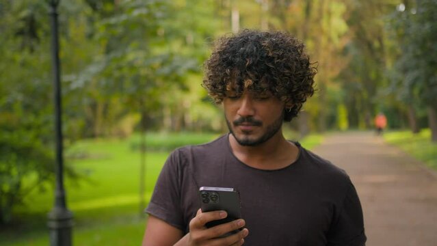 Arabian muslim Indian guy walking with smartphone in park ethnic thirsty man drinking water from sport bottle morning running outdoors happy smiling male check workout goals in mobile phone online app