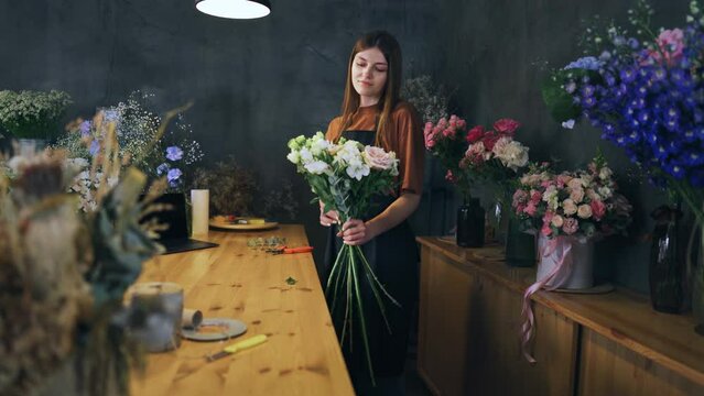 A woman is standing at a counter in a flower shop holding a bouquet of flowers. A bouquet designer creates a beautiful arrangement for customers. Portrait of a small business owner - a flower shop.