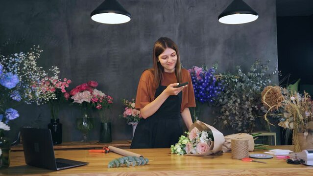 A woman stands at a table in a flower shop and takes a photo of a finished bouquet using her phone. A florist blogger takes photos of his work for social networks and the website.