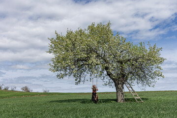 Young girl swinging on a swing in a pear tree. Wooden swing on which a happy woman swings outdoors. Swinging on a swing, dreaming of flying. Travel in nature in spring and summer. Healthy lifestyle.