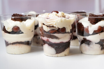 Cream and biscuit layered dessert. Homemade chocolate, vanilla cream and cherry trifles in transparent glasses, variation of dessert on white table
