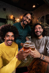 Vertical portrait of multiracial group of three men friends looking at camera while being in a bar toasting with beers, celebrating together and laughing.