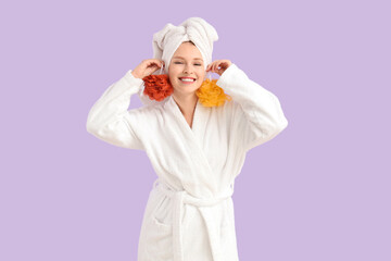 Young woman in bathrobe with loofahs after shower on lilac background