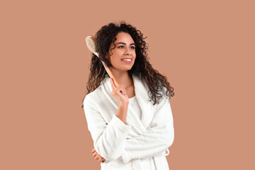 Young African-American woman in bathrobe with massage brush on brown background