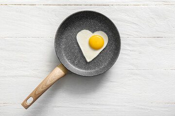 Frying pan with tasty fried egg in shape of heart on light wooden background