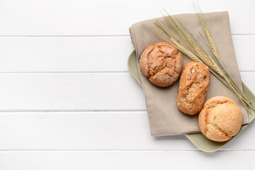 Plate with delicious buns and wheat ears on white wooden background
