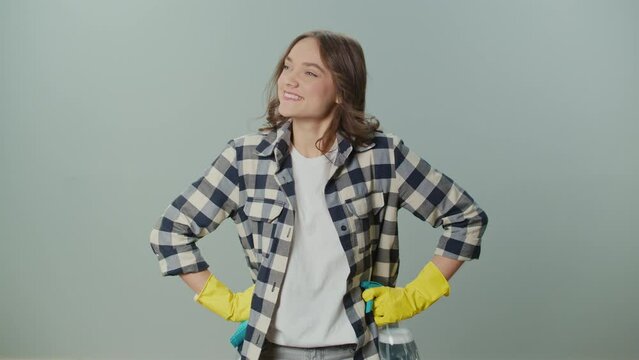 A Portrait of a Smiling Young Woman in Yellow Gloves Holding a Cleaning Spray Bottle and Rag, Satisfied with the Result of Her Cleaning on the Gray Background.Multi-tasking Cleaning Routines.