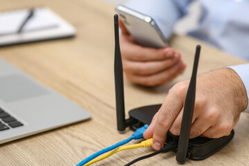 Man with smartphone and laptop connecting to internet via Wi-Fi router at wooden table, closeup