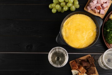 Fondue pot with melted cheese and different products on black wooden table, flat lay. Space for text