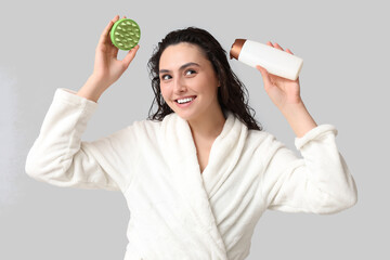 Happy young woman with hair scalp massager and bottle of shampoo on grey background