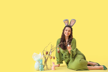 Beautiful woman in bunny ears with tree branches, Easter eggs and toy rabbits on yellow background