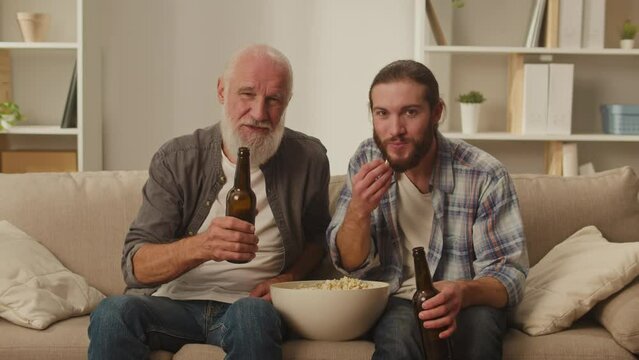 Senior and young friend on the sofa, with popcorn in their hands, involved in live movie watching, generational communication, pleasant family moments, watching together