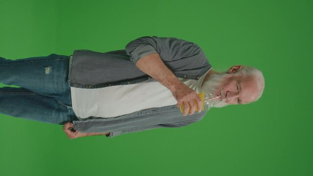Vertical View.Green Screen. A Smiling Old Man Drinks Orange Cocktail with Juice. An Elderly Man Feels Thirsty, Drinks Cold Juice. Healthy Lifestyle.