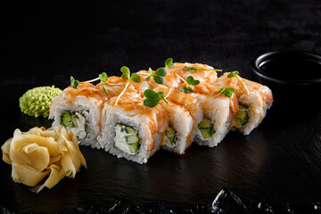 Shrimp roll with shrimp on top and cucumber and cheese inside, on a black slate with soy sauce, wasabi, and ginger