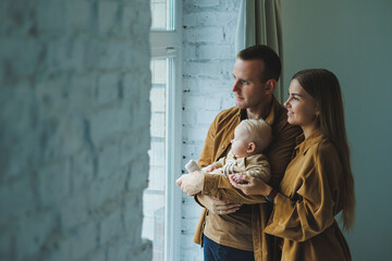 A happy mother and father hold their 4-month-old son in their arms and look out the window of their...