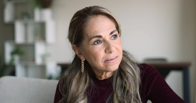 Close up shot serious pensive Latin mature woman deep in thoughts sit on sofa looking away, thinking, pondering spend weekend alone at home. Nostalgic mood, daydreams, midlife, concerns and disorders