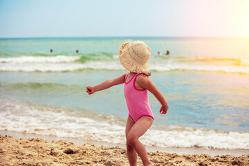 Happy little girl running on the beach in summer. The girl looks back. Kid in a straw hat