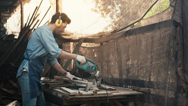 Handsome young craftsman wearing safety glasses and hearing protection headphones is using a wood saw to cut pieces, Carpenter or furniture designer is working in a workshop.	