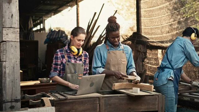 Diverse Craftspeople Working Together in a Woodworking Workshop Using Laptop and Hand ToolsDiverse Craftspeople Working Together in a Woodworking Workshop Using Laptop and Hand Tools