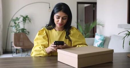 Indian retailer woman sit at desk prepare parcel box for sending to client, friend or family using modern mobile phone application, ordering express deliver e-services. Quick delivery services usage