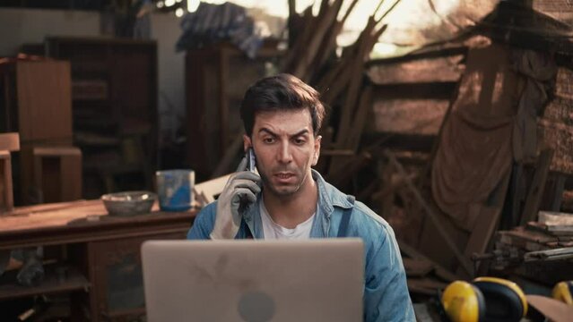 Handsome young craftsman was talking about his woodworking business over the phone, Carpenter is explaining the needs of a customer and is reading drawings with a laptop computer