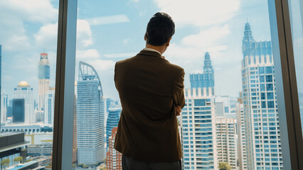 Fototapeta na wymiar Back view ambitious businessman standing in ornamented office gazing out window to cityscape skyline. Determination and business ambition drive business career toward to bright future