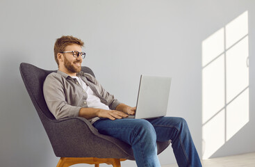 Happy, relaxed young man in casual shirt, jeans and eyeglasses sitting in comfortable armchair by...