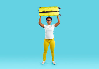 Full length portrait of afroamerican happy woman with yellow suitcase over her head smiling on turquoise background. She is wearing yellow jeans and white t-shirt. Travelling, tourism, journey, trip.