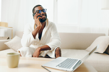 Smiling African American Man Working on Laptop in Cozy Home Office, Talking on Phone, Wearing...
