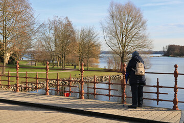alte Frau mit Rucksack und Gehstock steht allein auf einer Brücke und schaut auf Wasser