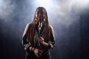 Waist up portrait of Black young woman with braided hair standing on stage with smoke background...