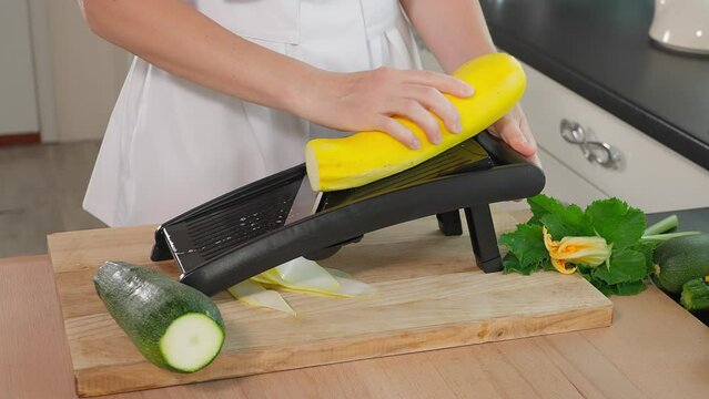 Chef's hands grate yellow zucchini slices on a mandalin grater, wooden table on a background of vegetables, home cooking, vegetarian food, vegetables rich in vitamins, diet. Close-up, side view