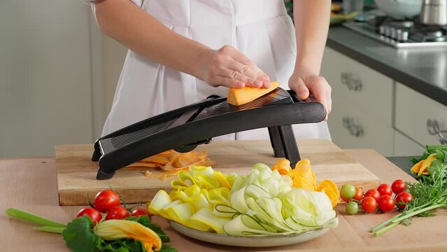 Chef's hands dangerously grate young carrots with slices on a mandalin grater, wooden table on a background of vegetables, home cooking, vegetarian food, vegetables rich in vitamins, diet. Close-up, s
