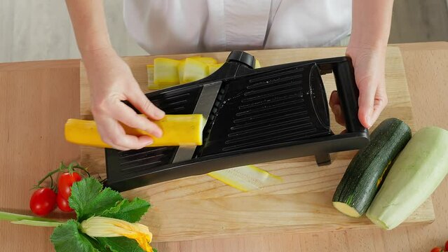 Chef's hands grate yellow zucchini slices on a mandalin grater, wooden table on a background of vegetables, home cooking, vegetarian food, vegetables rich in vitamins, diet. Close-up, top view