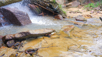 A Waterfall That Flows Into A Small, Clear River, With A Stretch Of Rocks At The Bottom