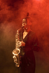 Vertical portrait of Black young woman playing saxophone on stage in nightclub with smoke effects