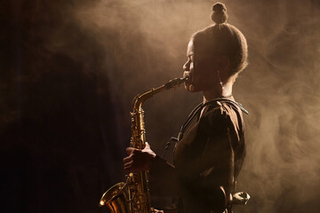 Ambient side view portrait of Black young woman performing jazz music on stage in dark nightclub and playing saxophone copy space - obrazy, fototapety, plakaty