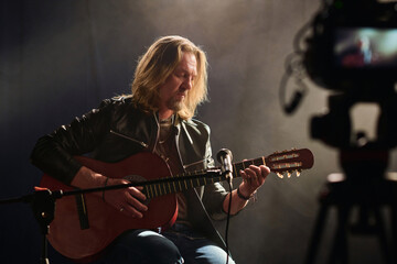 Portrait of long haired mature musician playing acoustic guitar with camera recording video copy...