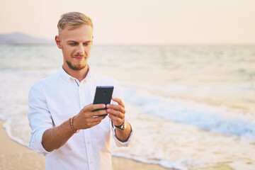 Vacation and technology. Portrait of young handsome man using smartphone on the sea shore.