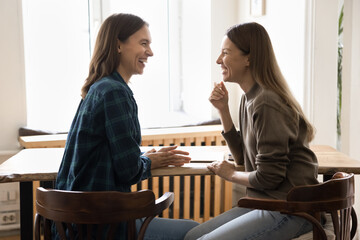 Happy young female business friends talking at workplace table, having fun, laughing, discussing funny creative ideas for project, enjoying office friendship, teamwork, cooperation