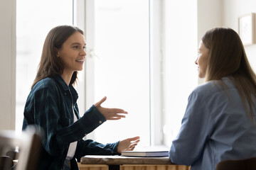 Happy positive millennial manager woman talking to female business partner at work table. Young...