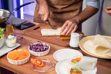 Cropped view of chef influencers presenting fresh salad roll on cooking step by step, streaming via smartphone on social media live channel, showing ingredients vegetable soft tortilla wrap. Sellable.
