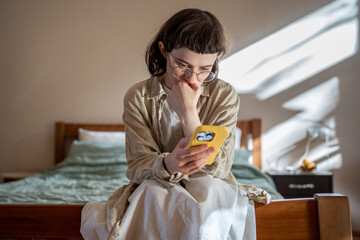 Pensive unhappy teenager sitting on bed with smartphone, scrolling web pages indifferently....