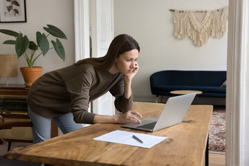 Serious focused beautiful freelance business woman working at laptop in home office, standing at...