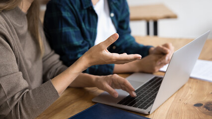 Young female coworkers brainstorming on project at laptop, talking at workplace table, using computer for discussion, moving hands, discussing work project. Cropped banner shot