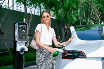 Young woman travel with EV electric car charging in green sustainable city outdoor garden in...