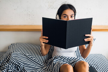 pretty sexy smiling woman sitting in bed in morning, listening to music on headphones and reading