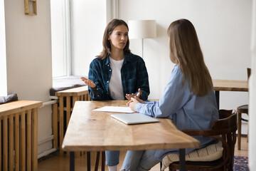 Two millennial business project partner women speaking at loft table, discussing work tasks,...